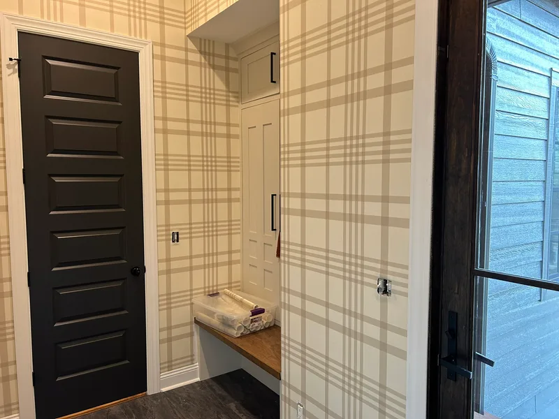 This mudroom features a classic plaid wallpaper in soft beige and cream tones, creating a warm, welcoming, and timeless atmosphere. The large-scale grid pattern adds subtle structure and visual interest while maintaining a light and airy feel that complements the white trim, dark door, and built-in cabinetry.