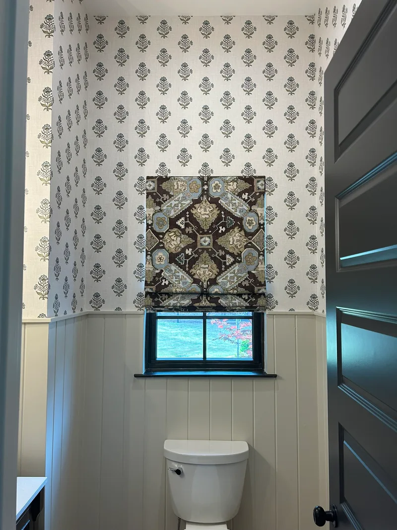 This elegant powder room features a sophisticated botanical wallpaper with a delicate repeating floral motif in soft neutral tones. The wallpaper covers the upper walls, creating a timeless and refined backdrop that pairs beautifully with the crisp white wainscoting below. A bold patterned Roman shade complements the wallpaper's design, adding depth and visual interest to the space.