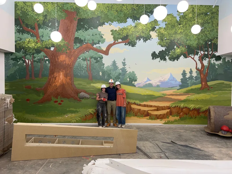This large 16' x 34' custom wallpaper mural transforms a new indoor playground at a church in Upstate, SC. The cheerful, illustrated forest scene features towering trees, rolling green hills, a winding path, distant mountains, and fun woodland details that create an immersive and joyful environment for kids. Professionally installed with seamless panel alignment across the expansive wall. Pictured here with the Charles Benton Wallpaper team after completion. Ready to bring creative, durable wall coverings to your church, business, or home? Contact us today!